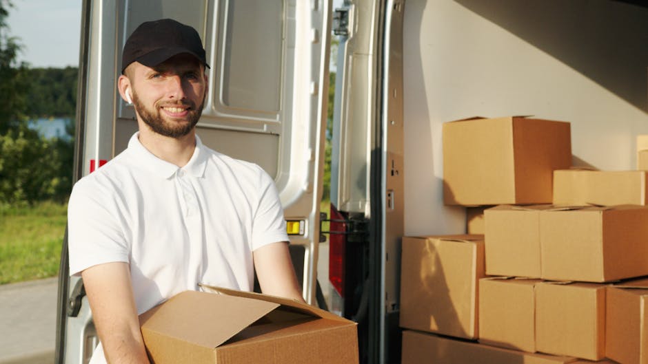 A man wearing a white polo shirt and a black cap is shown loading or unloading a cardboard box into or from the back of a moving van during a home relocation process. Inside the van, several other cardboard boxes are stacked neatly, some wrapped in plastic for protection. The background features green trees and a clear sky, indicating an outdoor setting near a residential area. The vehicle's interior is well-lit by natural light, highlighting the sturdy cardboard packaging used for safe furniture transport and packing during move logistics. The scene captures a professional move by Man with Van Eltham, with materials and equipment such as the boxes and the loading ramp visible, supporting house removals and furniture transport services.