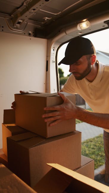 Two men are involved in a home relocation process, loading a large cardboard box onto a trolley in front of a white moving van parked on a brick driveway. One man, dressed in a blue jacket and black trousers, is inside the van, passing the box to a second man wearing a black puffer vest, grey hoodie, and dark jeans. The van's rear doors are open, revealing several packed cardboard boxes inside secure with labels, indicating an organized packing and moving process. Nearby, a black hand pallet truck is positioned on the driveway, ready for loading or unloading furniture or larger household items. The scene takes place outside a residential property with a brick wall, shrubbery, and a house with dark roofing visible in the background under cloudy lighting conditions. The image captures the organized logistics involved in furniture transport and professional removal services provided by Man with Van Eltham, supporting efficient and safe house removals.