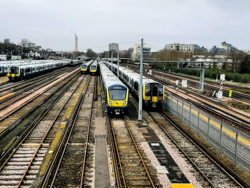 A collection of modern electric passenger trains parked on multiple railway tracks at a train station, with some trains facing forward and others aligned parallel. The trains are primarily silver with yellow and black accents, and their windows are visible on the sides. Overhead, there are electrical lines supported by metal poles running the length of the station. To the right, a tall metal fence separates the railway from adjacent areas. In the background, there are urban buildings, including residential and commercial structures, set against a cloudy sky. The station area is outdoor, with pathways and lighting posts, and the scene appears to be during daytime with overcast weather. This image reflects the logistics involved in home relocation and furniture transport, where careful planning and handling of transport vehicles, such as those managed by Man with Van Eltham, are essential for efficient moving services near Eltham Station and Mottingham Green, ELTHAM.