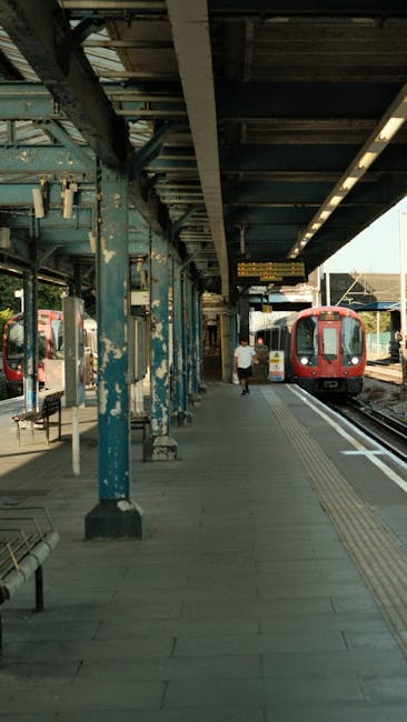 The image depicts a train station platform with an overhanging metal and concrete structure supported by weathered blue-painted steel columns. The platform is paved with concrete and features tactile paving for visually impaired passengers. A red train is stationary on the track to the right, with open doors indicating it may be preparing for departure or arrival. A person dressed in casual clothing is walking along the platform, heading towards the train. There are metal benches positioned along the platform, and additional tracks are visible in the background, along with station signage and a street scene partly visible through the open sides of the station. The scene is illuminated by natural daylight, casting soft shadows, and the overall setting reflects typical commuter station activities, with a focus on train travel and passenger movement, relevant to the context of UK train stations and transportation services.