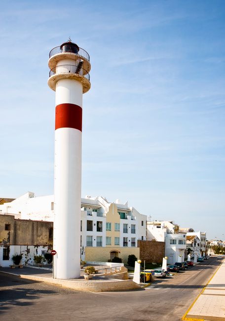 A tall, white lighthouse with a red horizontal stripe near the middle, situated on a paved area adjacent to a street. The lighthouse features a viewing platform with a metal railing and a small lighting apparatus at the top. In the background, there are modern white residential buildings with multiple balconies and flat roofs, lined up along the street. Several parked cars are visible along the roadside, and the sky above is clear with light clouds. The scene is well-lit, indicating daytime, and the environment appears clean and maintained. As part of a house removals or home relocation context, the image may represent a coastal location or guide for moving items near seaside landmarks, with Man with Van Eltham providing professional removals services in the area.